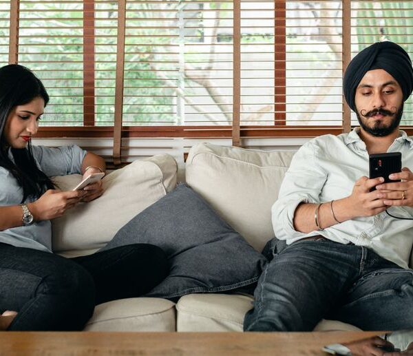 A husband and wife sit side by side on the couch, each absorbed in their phone instead of connecting with one another, reflecting emotional distance in the marriage.