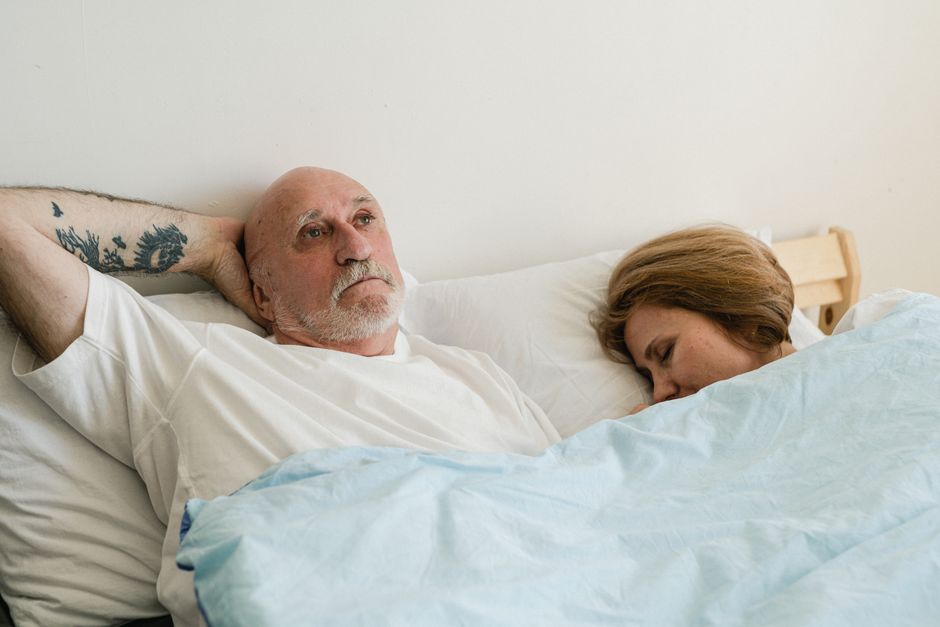A man lies awake deep in thought beside his sleeping partner, keeping his private reflections to himself.
