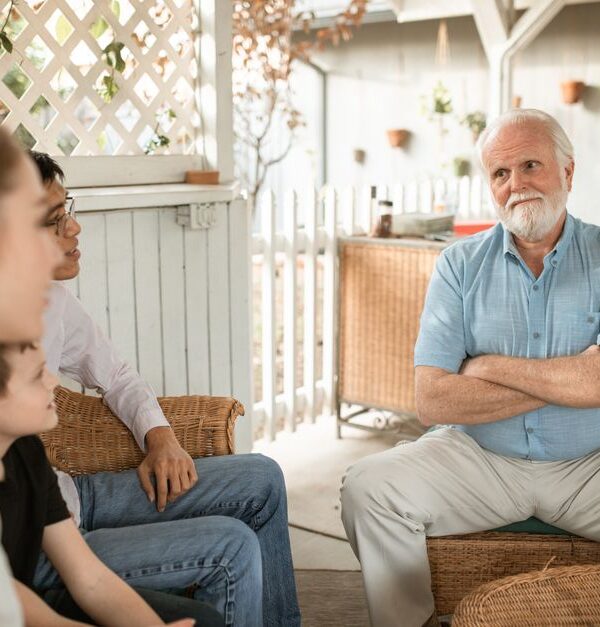 An older parent sits with crossed arms while his adult child's family gathers around, illustrating tension that can arise from saying the wrong things to adult children.