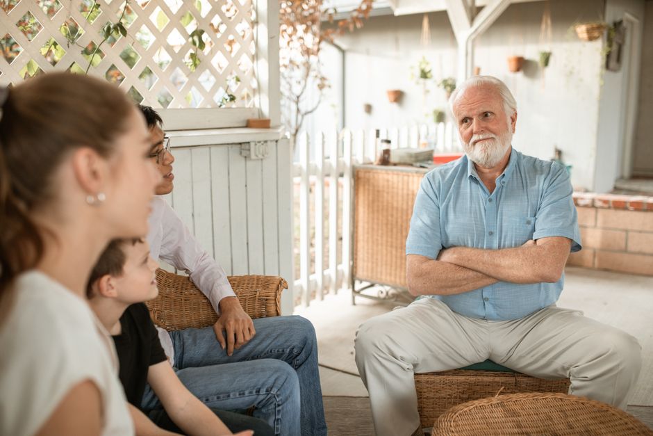 An older parent sits with crossed arms while his adult child's family gathers around, illustrating tension that can arise from saying the wrong things to adult children.