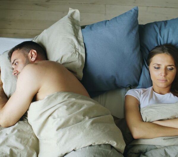 A couple lies in bed turned away from each other, the woman's guarded expression hinting at underlying mistrust.