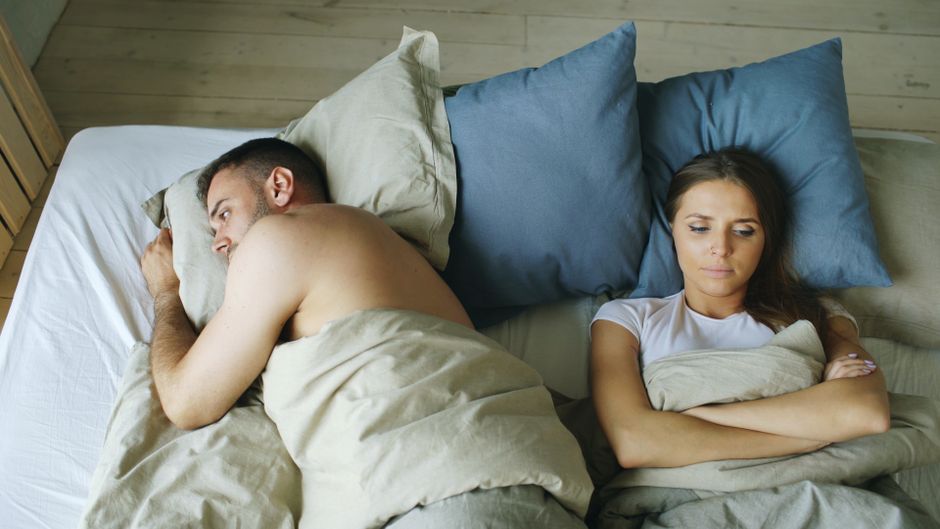 A couple lies in bed turned away from each other, the woman's guarded expression hinting at underlying mistrust.