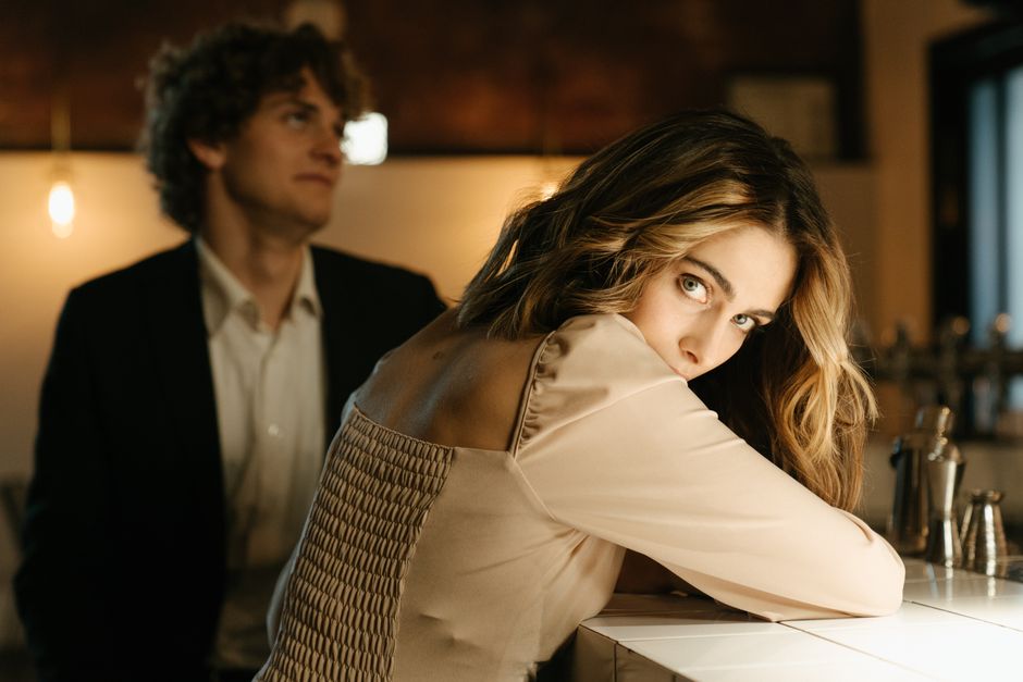 A woman at a bar glances back invitingly over her shoulder while a man nearby looks her way, hinting she wants him to approach.