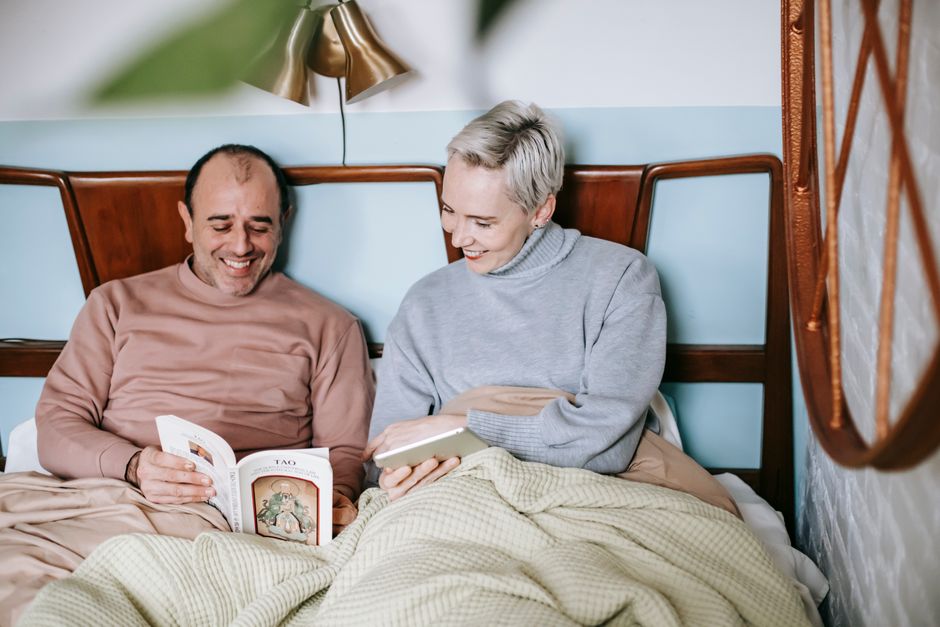 An older couple spends weekly quality time together, relaxing and reading in bed while smiling at each other.