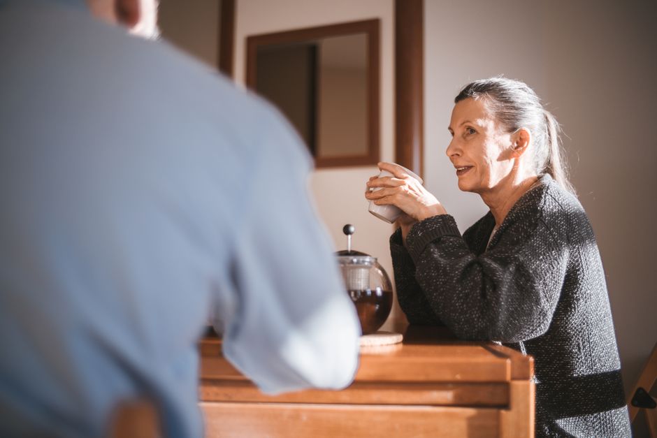 An older married woman discusses household matters with her husband over coffee in their kitchen.