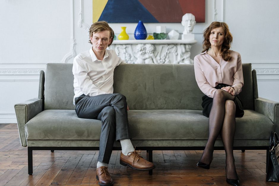 A couple sits apart on a couch, each facing forward separately as if mentally distanced from one another.