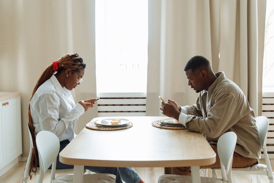 A couple sits at the same table but emotionally distant, each absorbed in their phone instead of sharing a meal together.