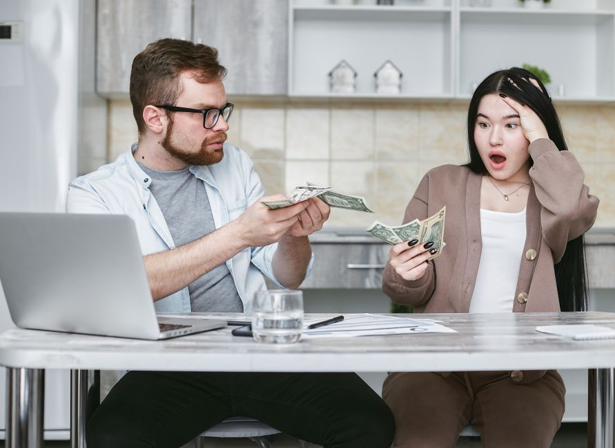 A man forcefully offers cash to a shocked woman at a table, symbolizing control over shared finances.