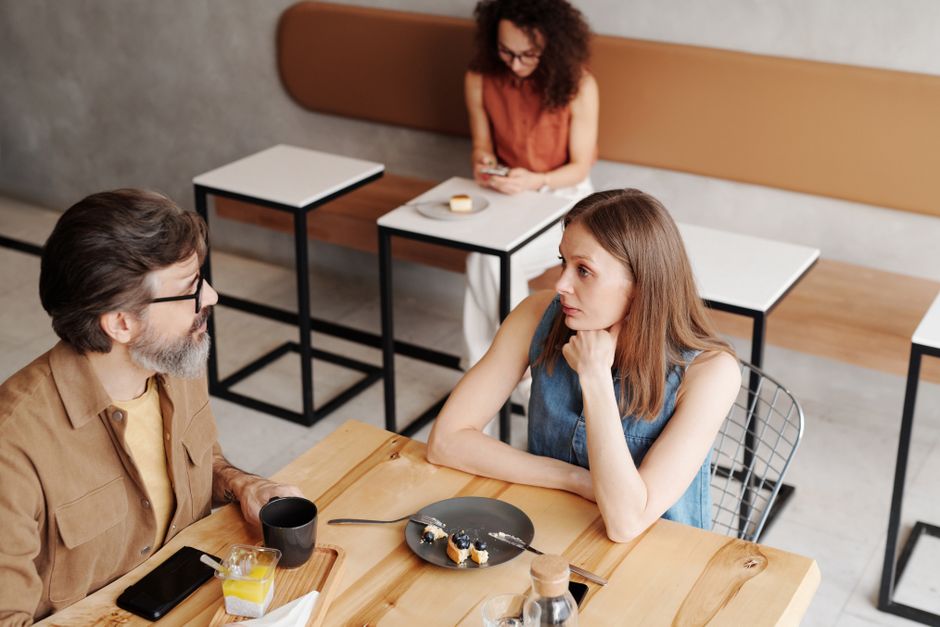 A couple sits at a cafe table with a distant, disconnected look, as if they've stopped truly checking in with each other.