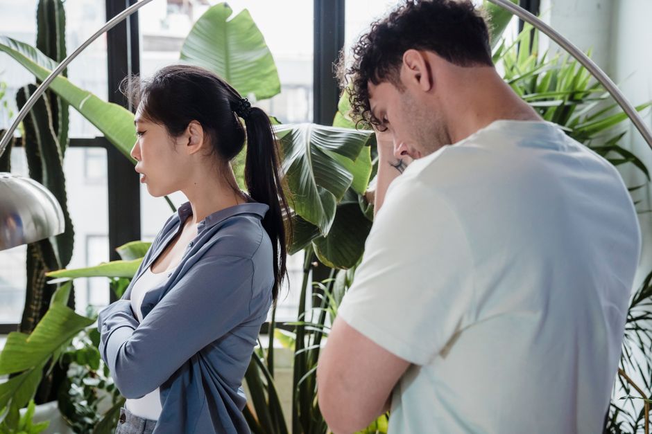 A couple stands back to back with crossed arms, avoiding each other's gaze, reflecting unresolved tension and a lack of apologies between them.