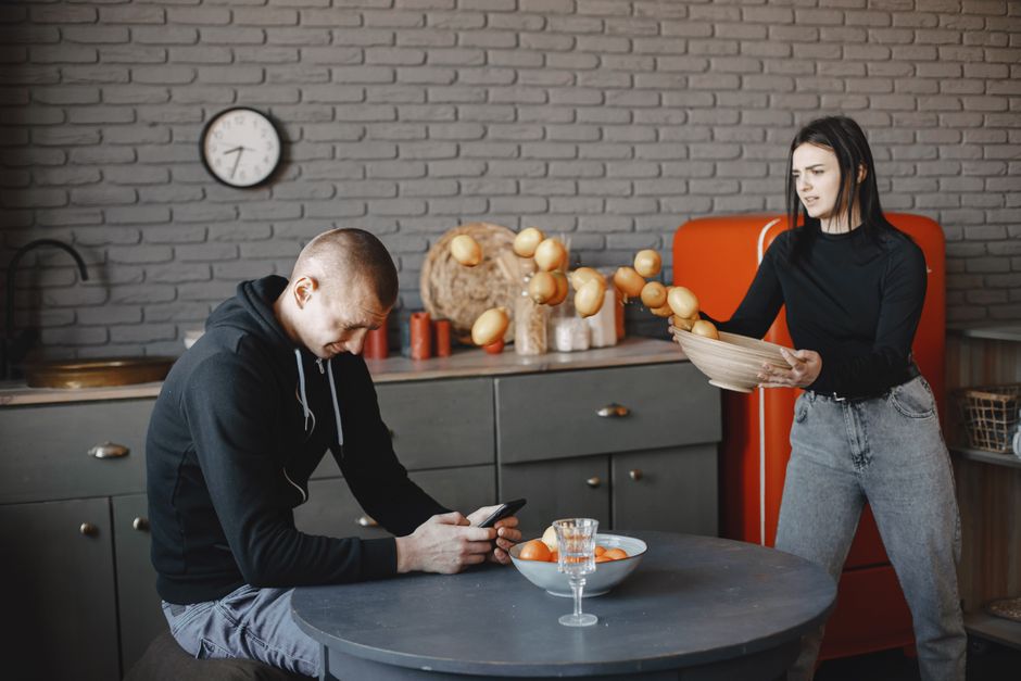 A woman watches suspiciously as her partner is absorbed in his phone, hinting at tension over privacy and trust in the relationship.