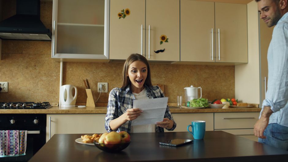 A woman reads surprising news on paper while her partner watches from across the kitchen, unaware of what she's just learned.