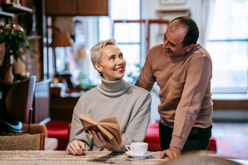 A husband stands over his wife with a polite smile, but their exchange feels oddly formal rather than warm.