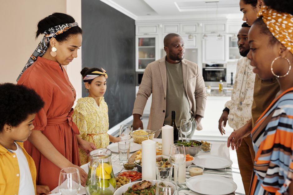 A man stands at a family gathering while others around the table seem to be pointing out something he did wrong in front of everyone.