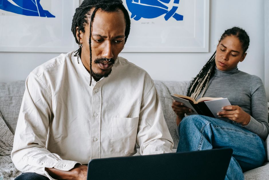 A husband absorbed in his laptop making his own plans while his wife reads alone beside him, disconnected from shared decisions.