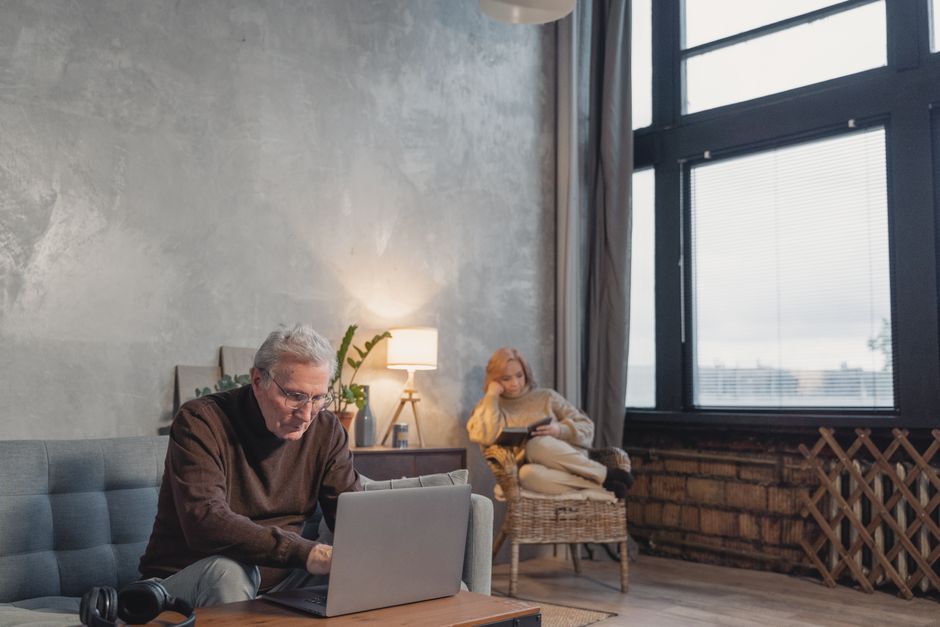 Husband sits apart on the couch absorbed in his laptop while his wife reads alone in a chair across the room, each in their own separate space.