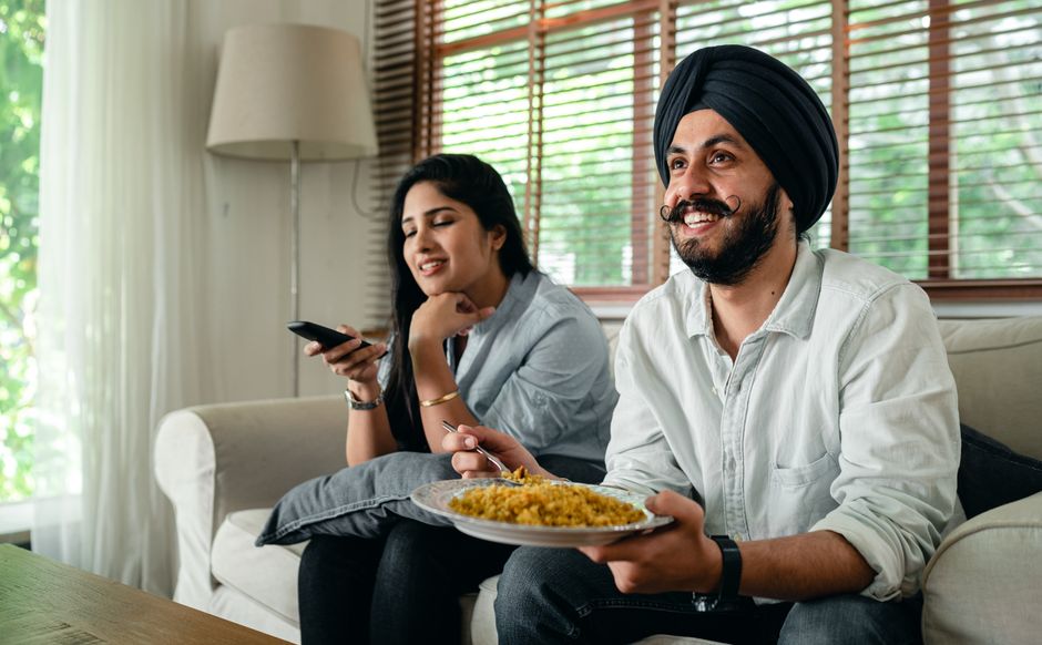 A husband eats his meal on the couch in front of the TV instead of joining his wife at the dinner table.