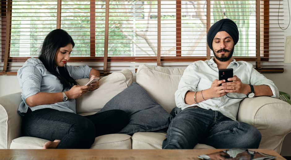 A man and woman sit apart on a couch, both absorbed in their phones instead of each other, reflecting the quiet distance men notice when a partner pulls away.