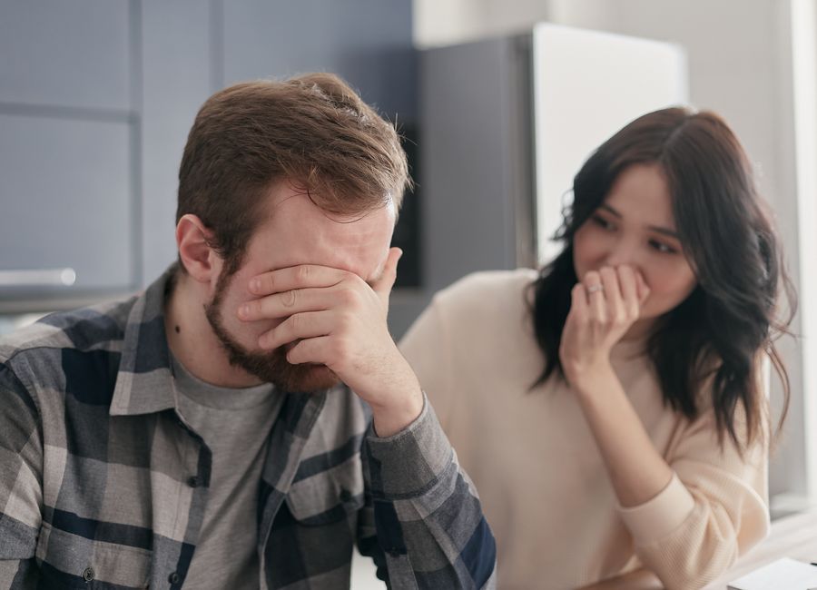A man looks embarrassed covering his face while a woman nearby reacts, illustrating the discomfort men feel when corrected in front of others.