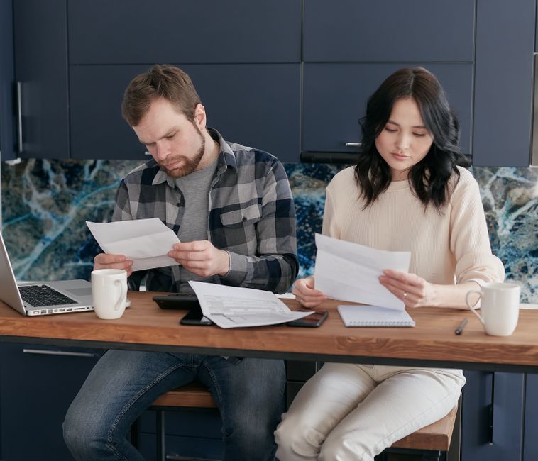 A husband and wife sit together but emotionally distant, each absorbed in their own paperwork without exchanging a word or glance.