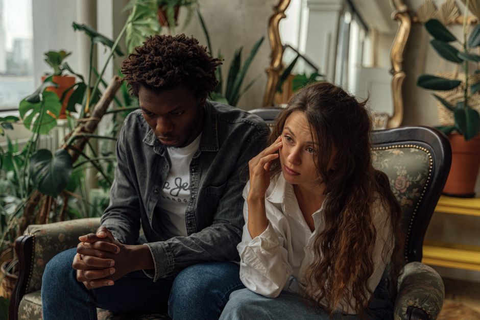 A couple sits in heavy silence on the couch, no laughter or connection between them, reflecting a husband emotionally checked out of the marriage.
