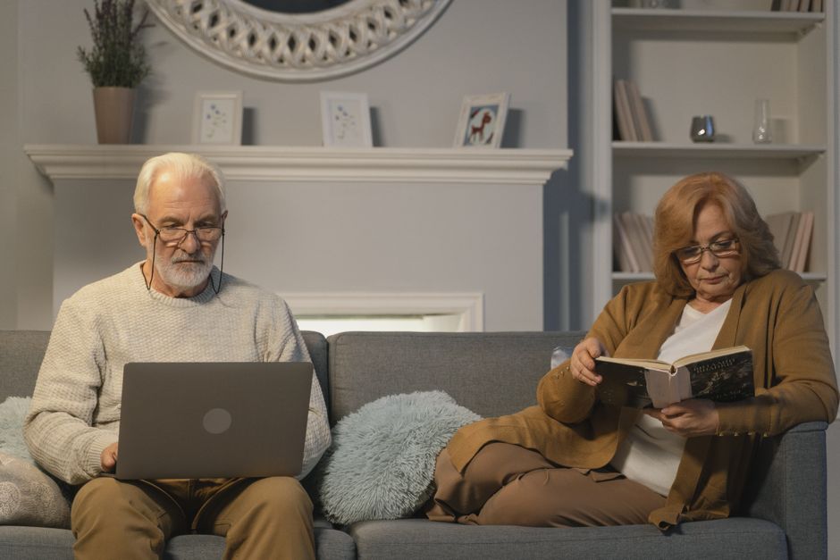 An older husband buried in his laptop while his wife reads alone, both disconnected and not sharing small moments of news with each other.