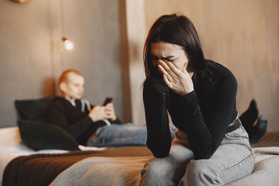 A woman tearfully reacts to harsh words while the man remains distant, scrolling his phone nearby.