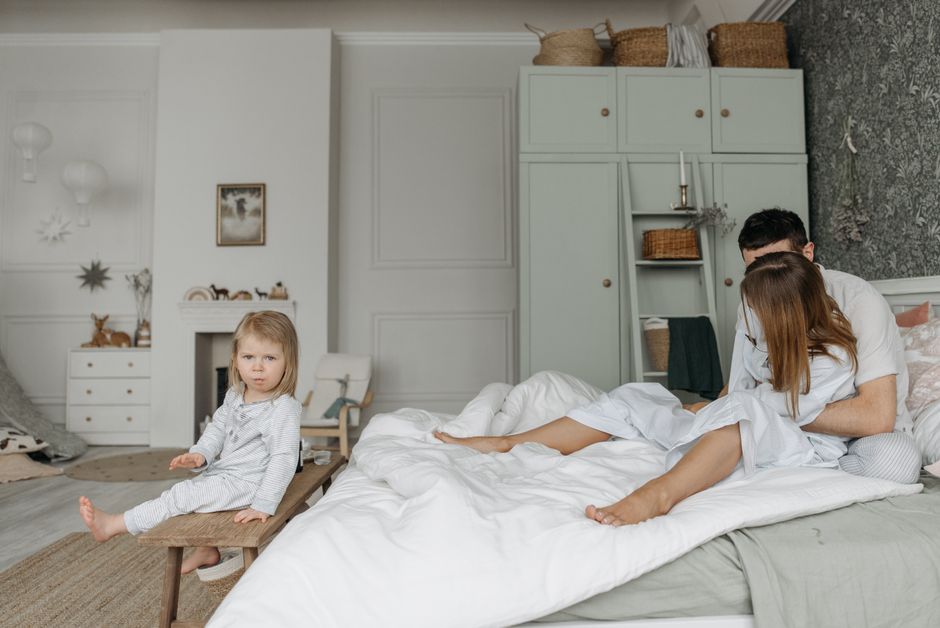 A father and mother share an intimate moment on the bed while their child sits alone nearby, illustrating feeling second to the children's needs.
