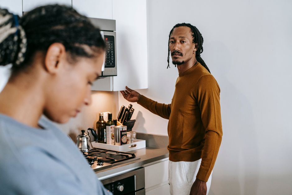 A husband looks checked out and indifferent while his wife tries to talk to him in the kitchen.