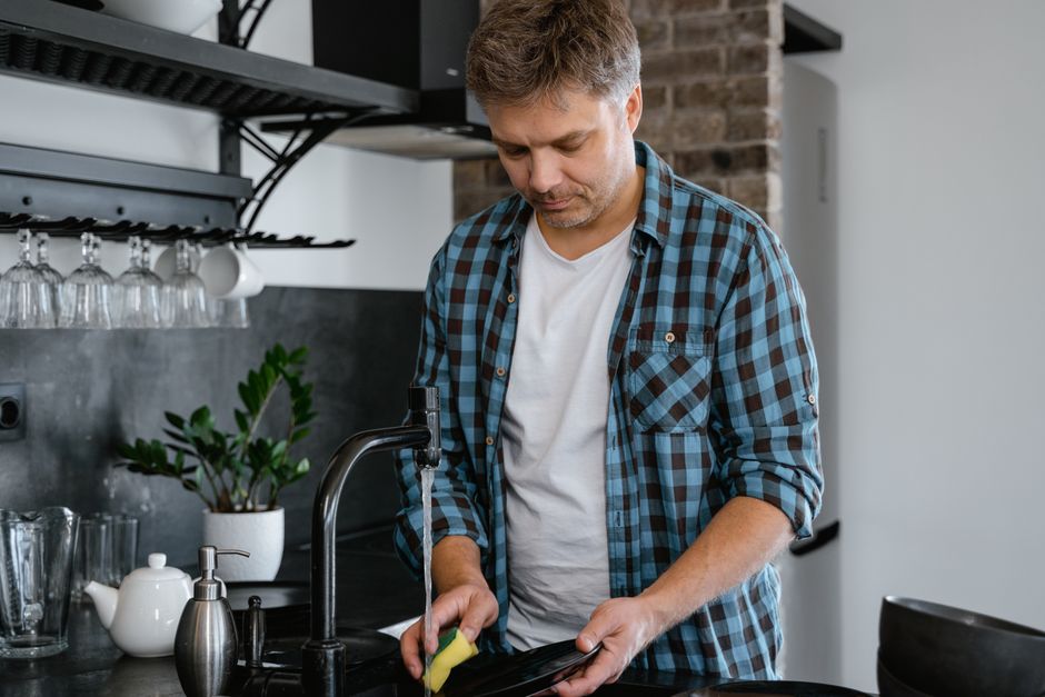 A man quietly washes dishes alone in the kitchen, his small, unnoticed effort going unthanked.