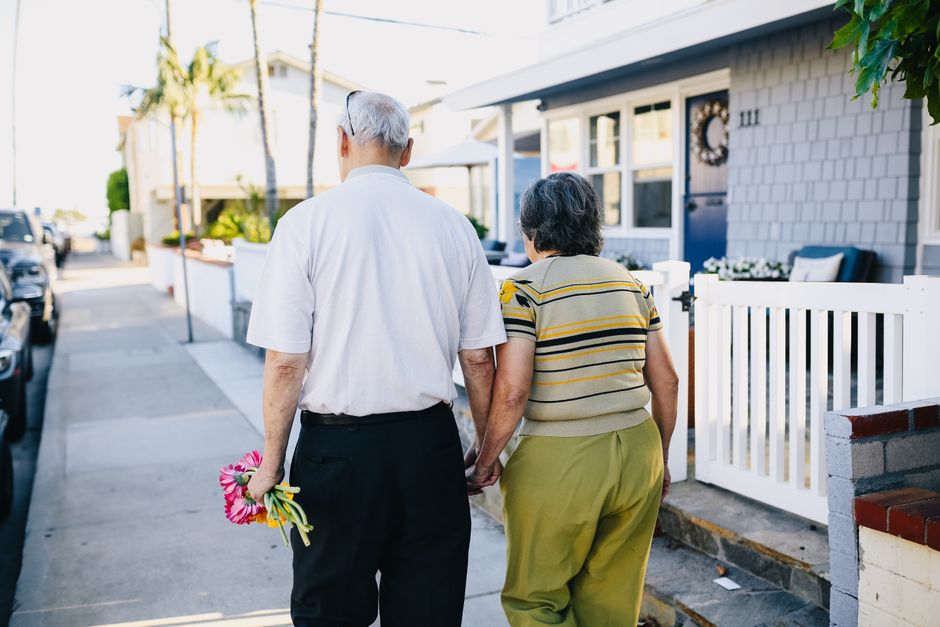 An elderly couple walks hand in hand down the street, the man carrying flowers, symbolizing a lifelong commitment to grow old together.