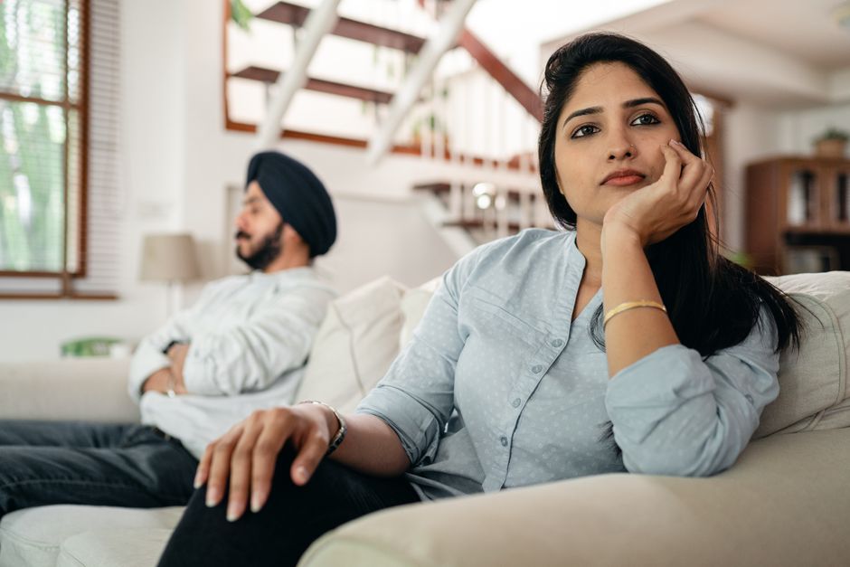 A woman looks thoughtfully distant from her partner on the couch, reflecting the quiet moments when he wishes she'd reach out first.