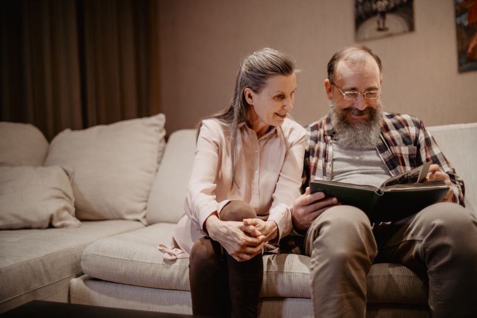 A husband and wife sit close together on the sofa, smiling as they look through an old photo album full of memories.