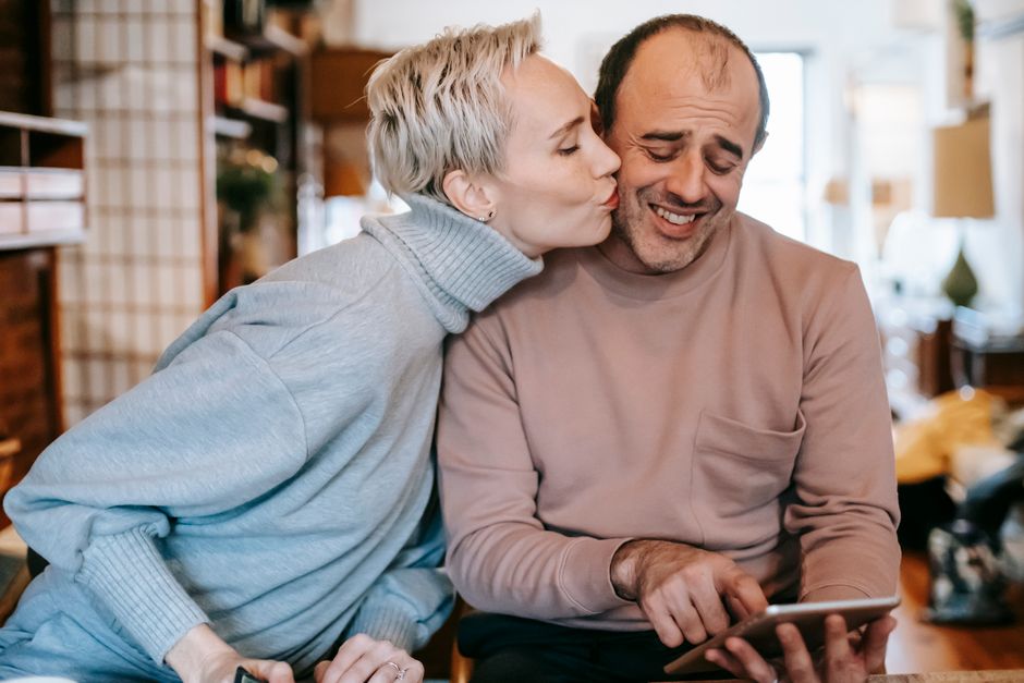 A wife kisses her husband's cheek as he reads a surprise note on a tablet at home.