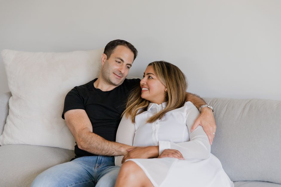 A husband and wife sit side by side on the couch, smiling and embracing each other closely.