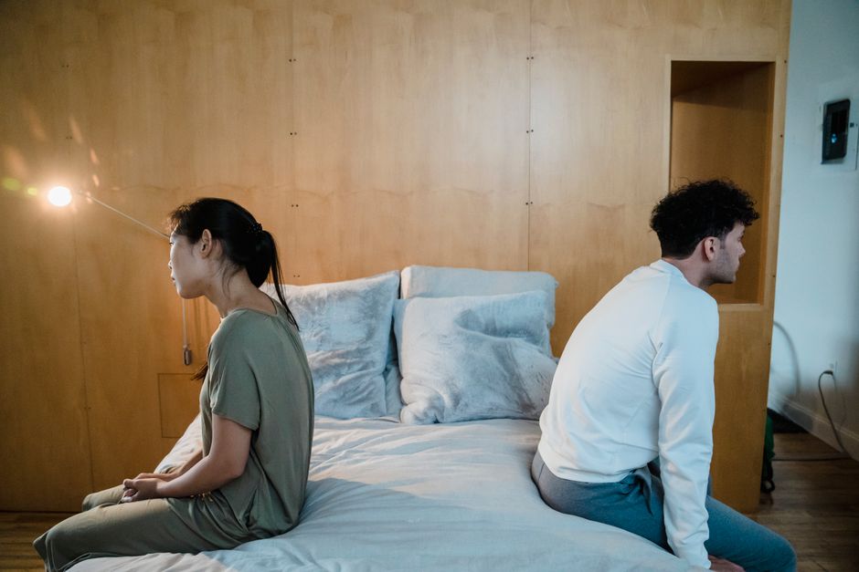 A couple sits back-to-back on the edge of a bed, emotionally distant from one another.