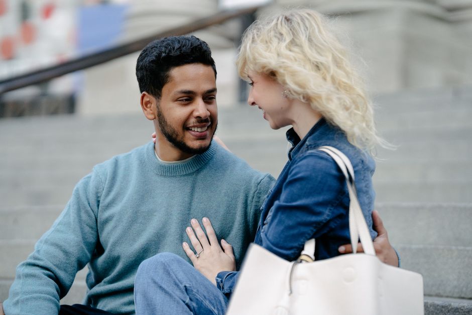 A man smiles bashfully while sitting close to a woman on outdoor steps, showing a subtle sign he likes her.