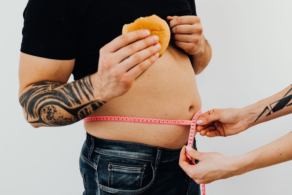 A man holding a burger gets his waist measured, as if shrinking the truth a little more each time.