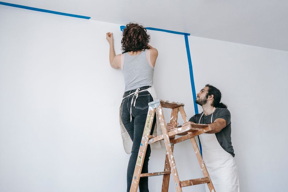 A man helps steady the ladder and assists with home repairs while his partner works on painting the wall.