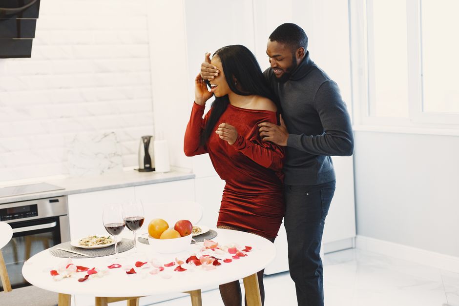 A man surprises his smiling girlfriend with a planned romantic evening, complete with a decorated table and rose petals.
