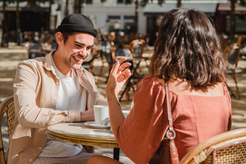 A man smiles warmly while talking closely with a woman over coffee at an outdoor cafe, hinting at a flirty conversation.