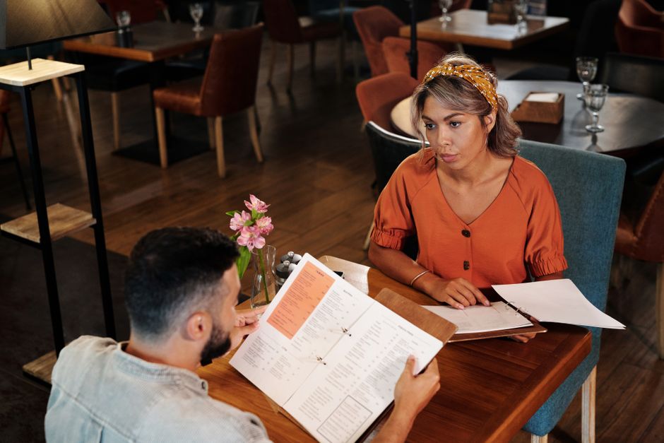 A man and woman review a restaurant menu together, her questioning look hinting he already knows what he wants but isn't saying.