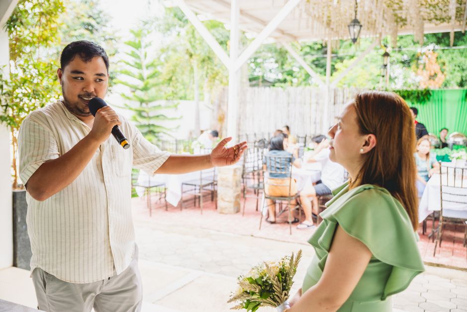 A man publicly speaks and gestures warmly toward a woman, evoking the idea of a man speaking well of her in front of others.