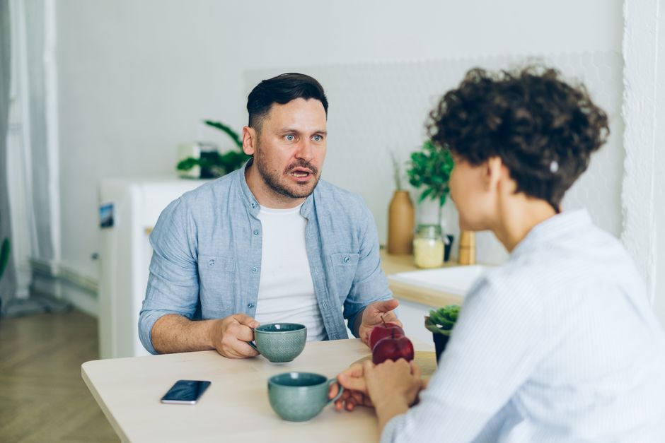 A couple having a tense conversation over coffee, the man explaining himself while the woman looks skeptical.