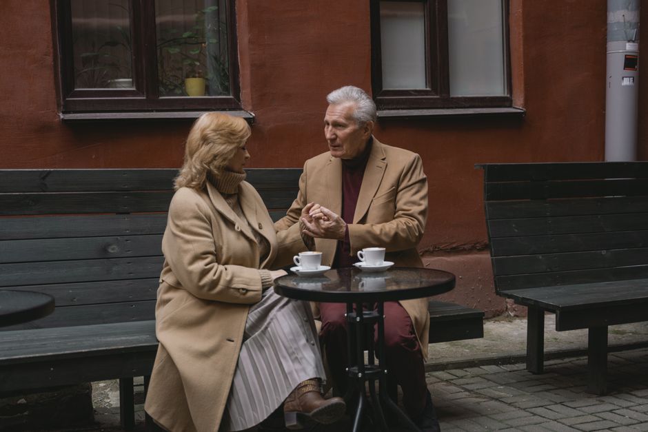An older man and woman share a tender moment over coffee, holding hands at an outdoor café table, reflecting a loyal, decades-long bond.