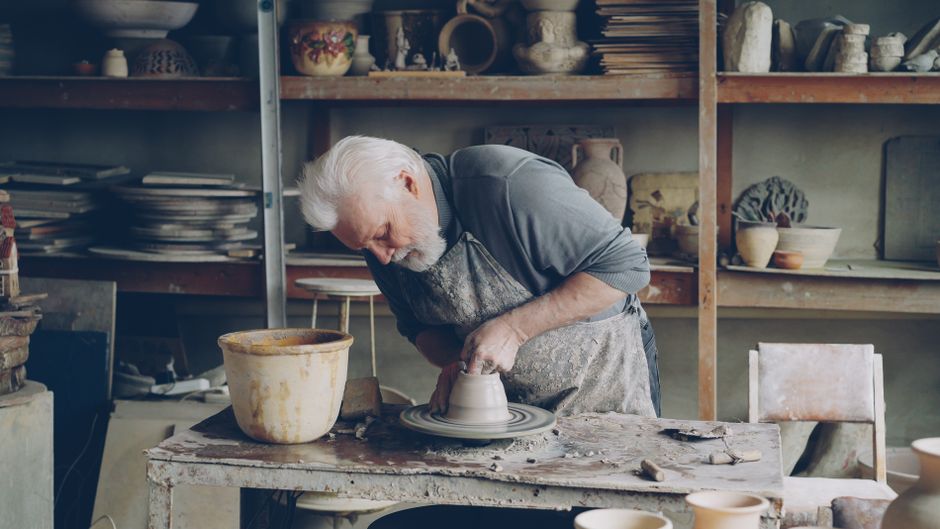 An older man enjoys his hobby of pottery making, shaping clay on a wheel in his own workshop.