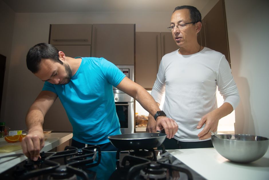 An older man teaches a younger man how to cook breakfast together at the stove.