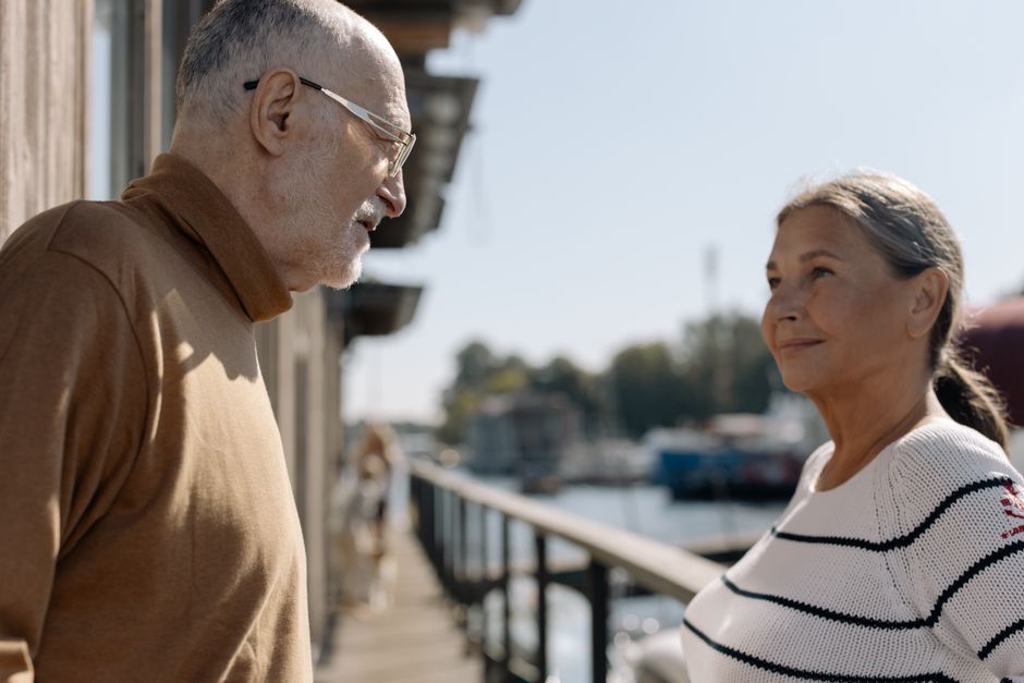 An older man gazes attentively at his wife as she speaks to him on a sunny dockside walk.