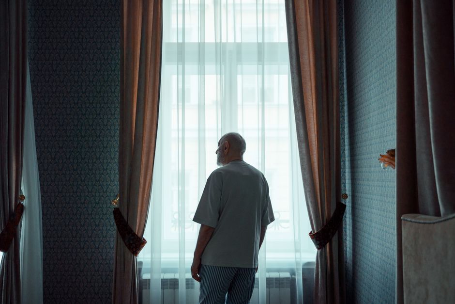 An older man stands quietly by a window, reflecting during a difficult moment, choosing to stay rather than walk away.