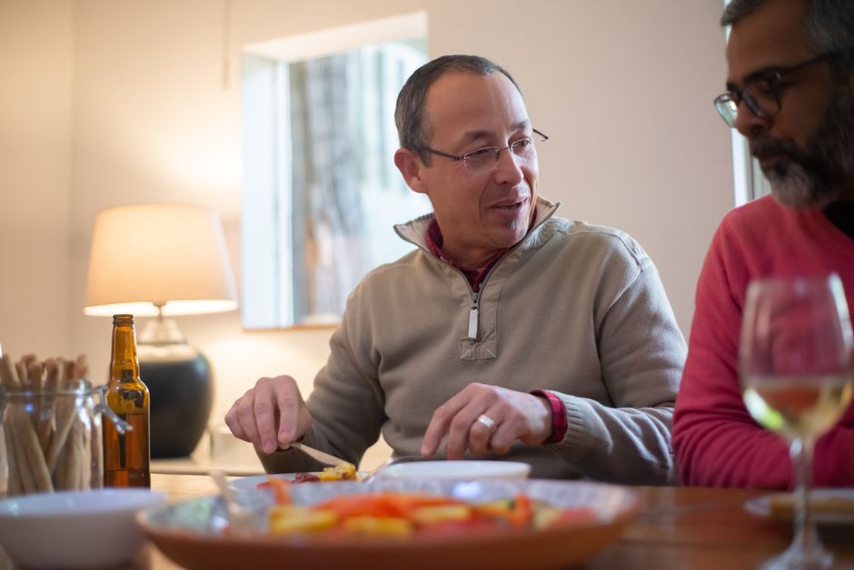 A mature man enjoys dinner conversation at the table with a friend, embodying the habit of eating dinner together.